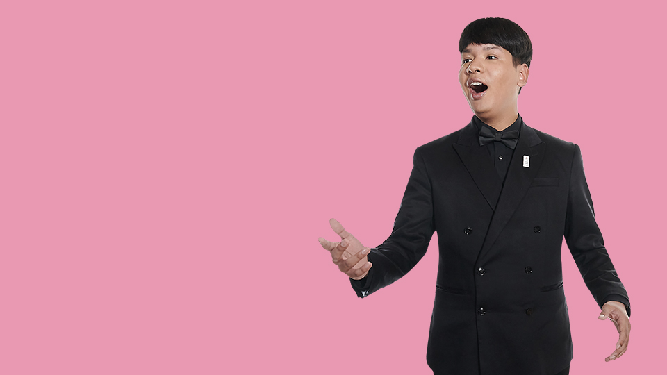 A man wearing a black double-sided blazer, with short hair, singing, with a light pink background.